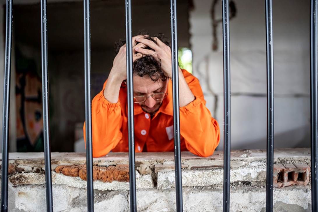 A despondent man in an orange jumpsuit is behind bars, head in hands. The image emphasizes the distress associated with potential false imprisonment and detention.